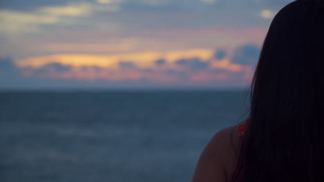 Back-view-of-an-unrecognizable-woman-admiring-a-spectacular-sunset-standing-by-the-sea-and-touching-setting-right-hair-which-is-blowing-on-breeze-wind