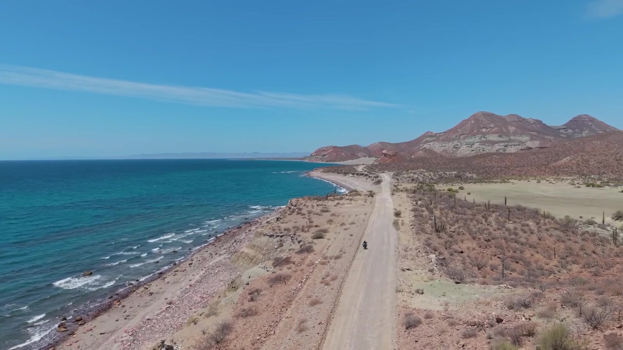 Uma ampla tomada aérea de acompanhamento da estrada de terra acidentada e solitária que serpenteia pelos penhascos áridos da costa da Baja Califórnia
