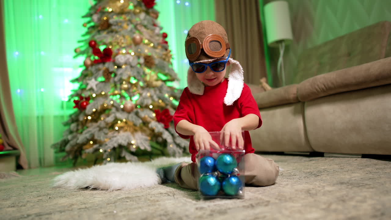 Caucasian toddler wearing aviator cap and sunglasses holds a box of toys. Child sits down on the floor to play with toys. Christmas tree at backdrop.