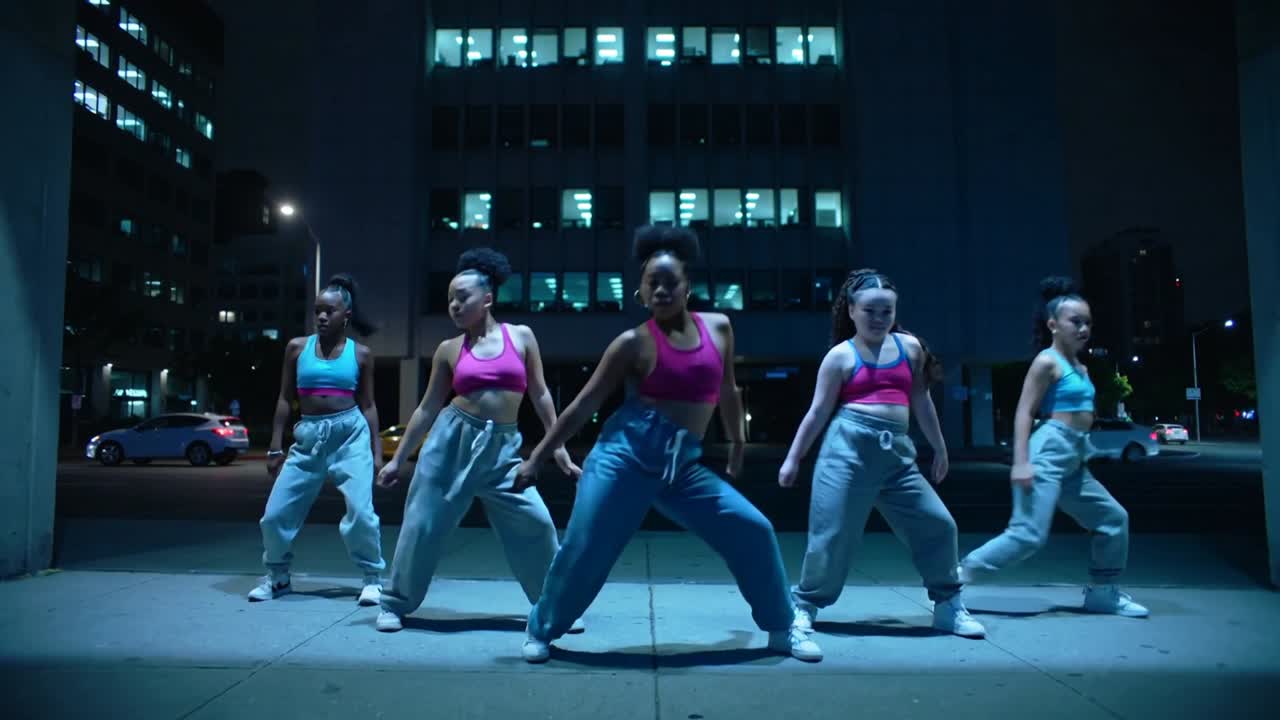 Vijf dansers beginnen op de maat en voeren hippe moves uit op de stoep, schuivend en eindigend in een squat, in croptops.