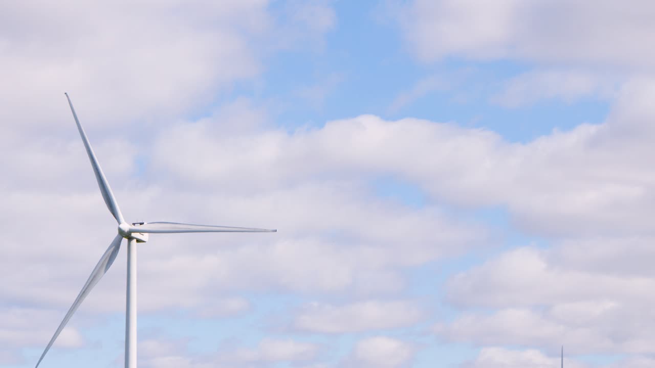 A single wind turbine slowly rotates in a wide shot, set against a bright blue sky with scattered clouds. Daylight, steady camera, tranquil mood