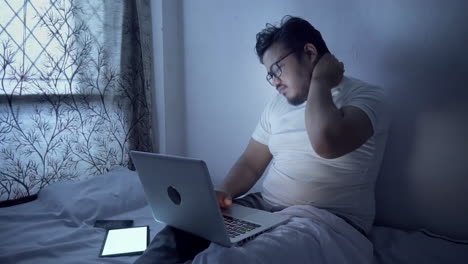 indian young man using laptop and overworking at night on bed, having pain in neck