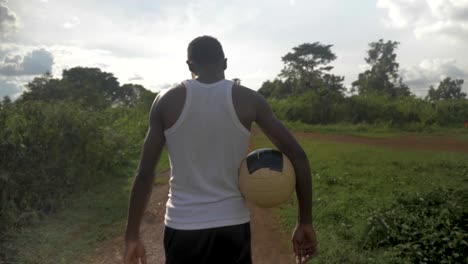 a slow motion tracking shot from behind of a young african man holding a football while walking towards the sun