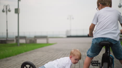 a little boy is riding his bicycle along a paved path when he falls while attempting to maintain balance, his older brother rides behind him, coming closer