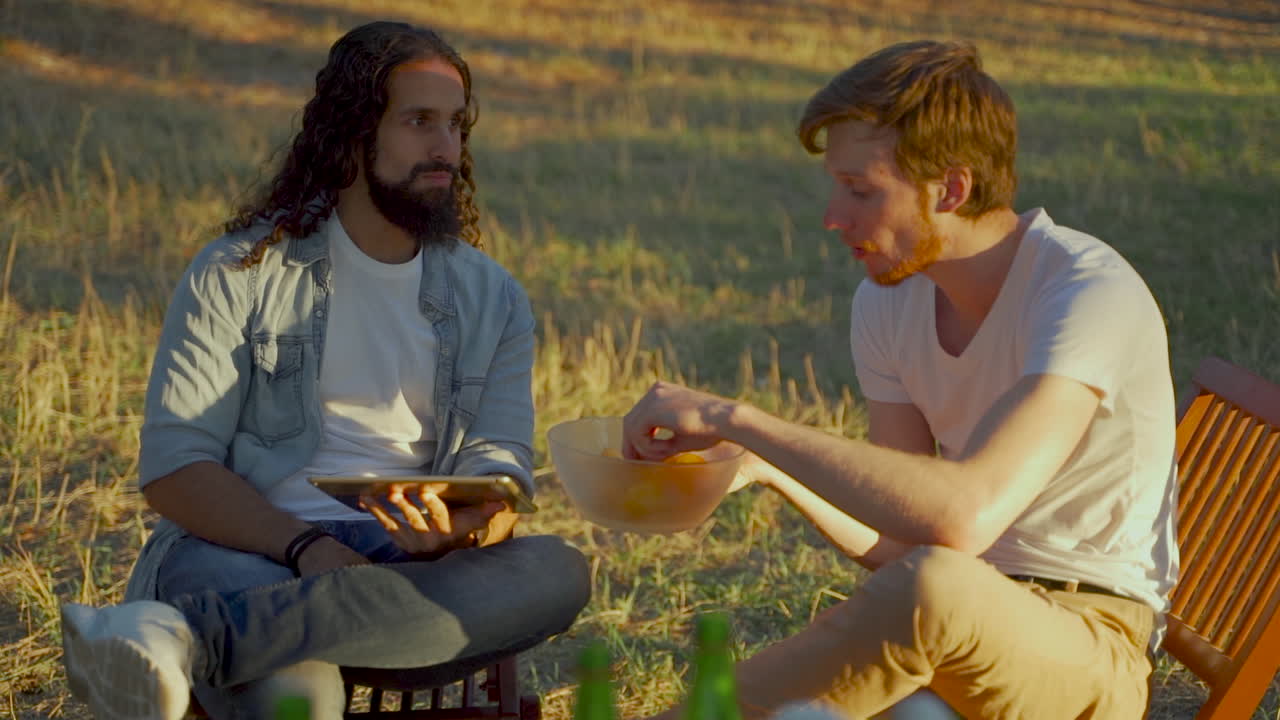 hombres jóvenes con una tableta y comida hablando en el campo. gente disfrutando de un picnic en la naturaleza.