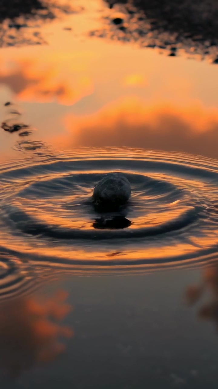 Vertical video: Dropping tiny water droplet forming crown round ripples on mirror puddle at sunset