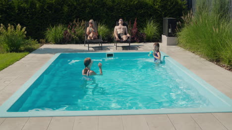 la familia se relaja en su piscina en el patio trasero de la casa. los adultos toman el sol en sillas de sol, dos niños nadan en la piscina