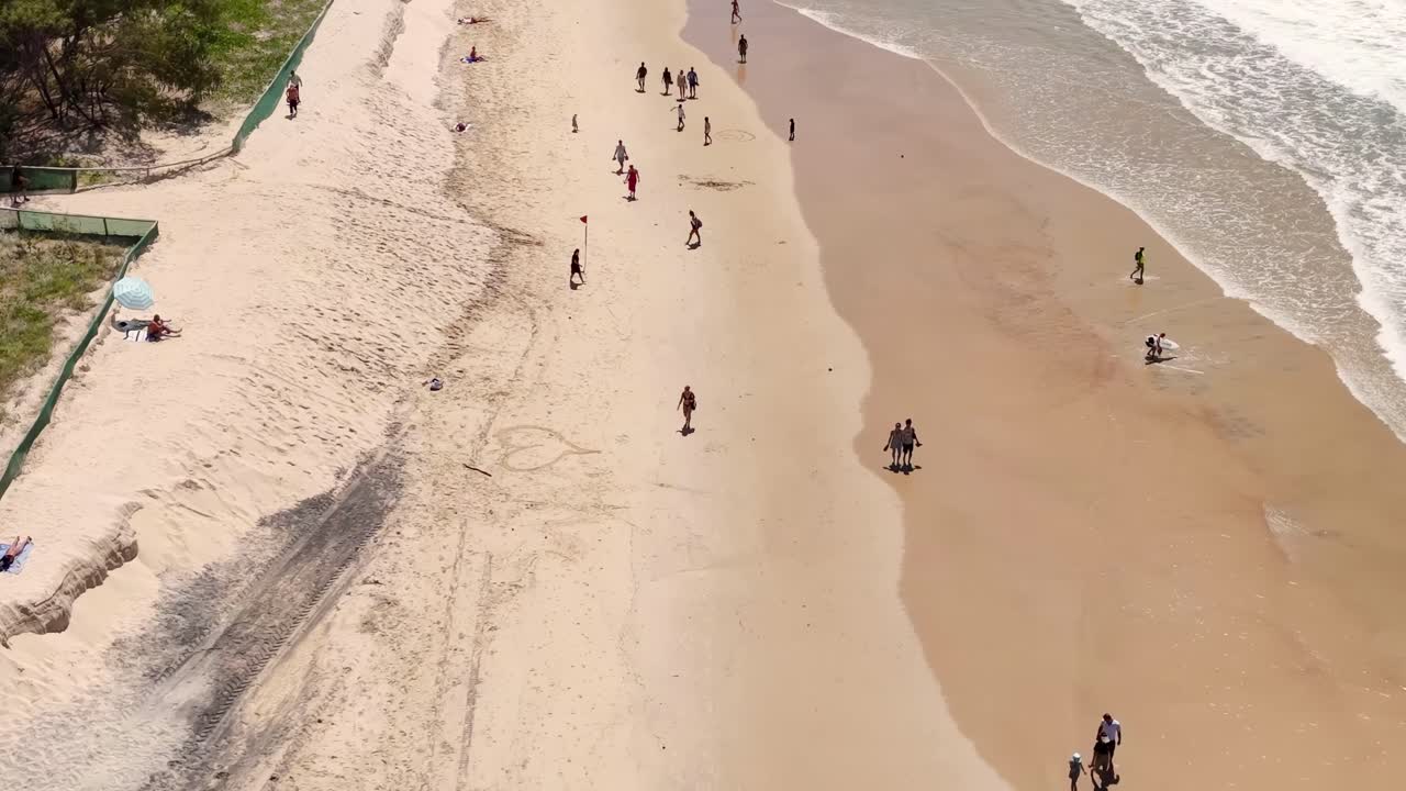Drone footage shows people walking along a sandy beach with gentle ocean waves lapping the shore.