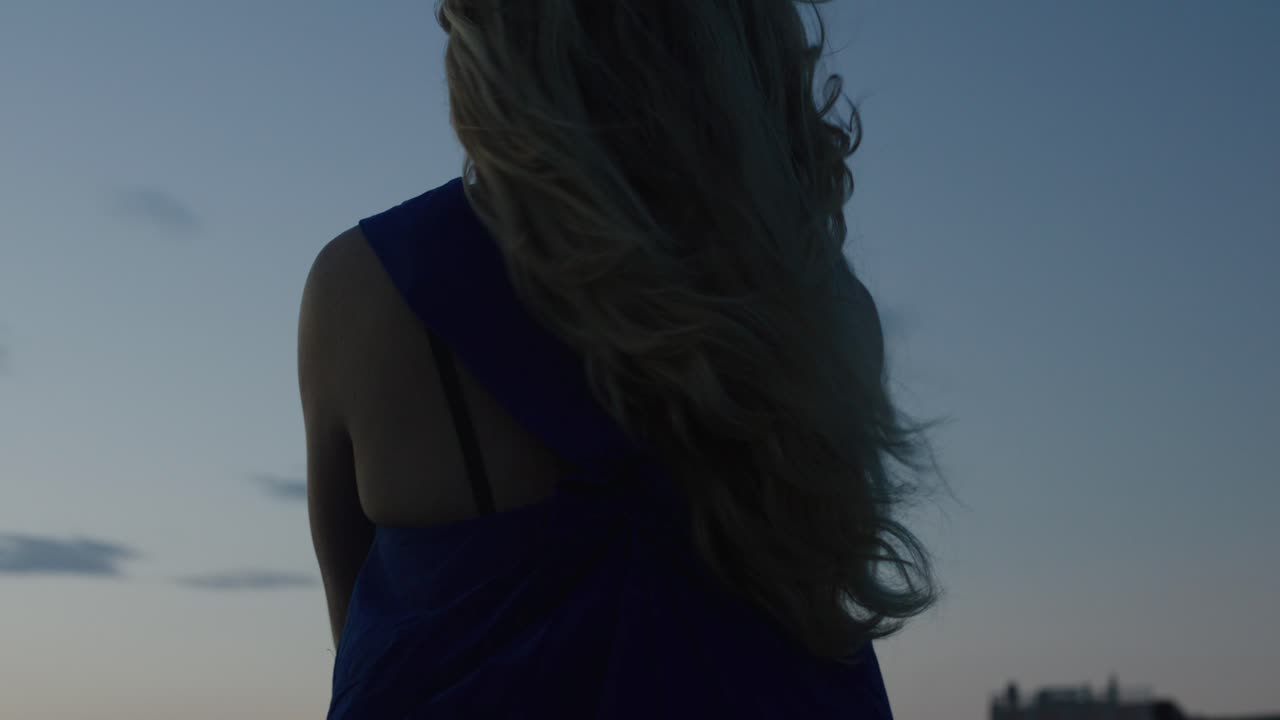 Blonde haired woman sitting on a ledge in deep thought as wind blows enjoying the sunset in Oia, Santorini, Greece Europe