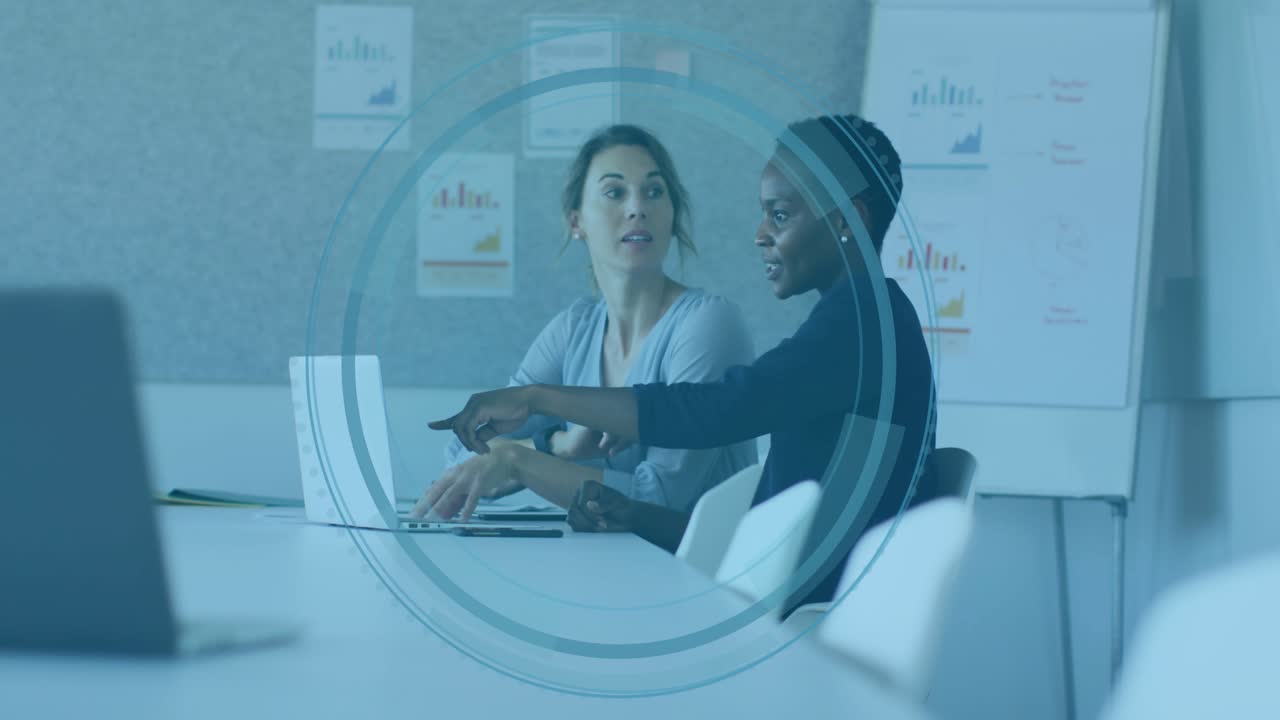 Two women at table right woman pointing laptop causing circle over them showing business charts