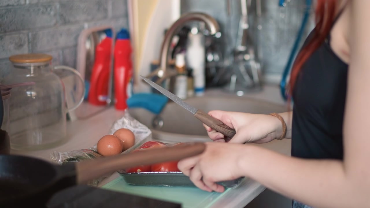 Giovane donna prepara il pasto mattutino. Donna dai capelli rossi taglia attentamente la salsiccia al lavello. Giovane donna con vivaci capelli rossi prepara abilmente l'essenziale per la colazione in un'accogliente cucina.