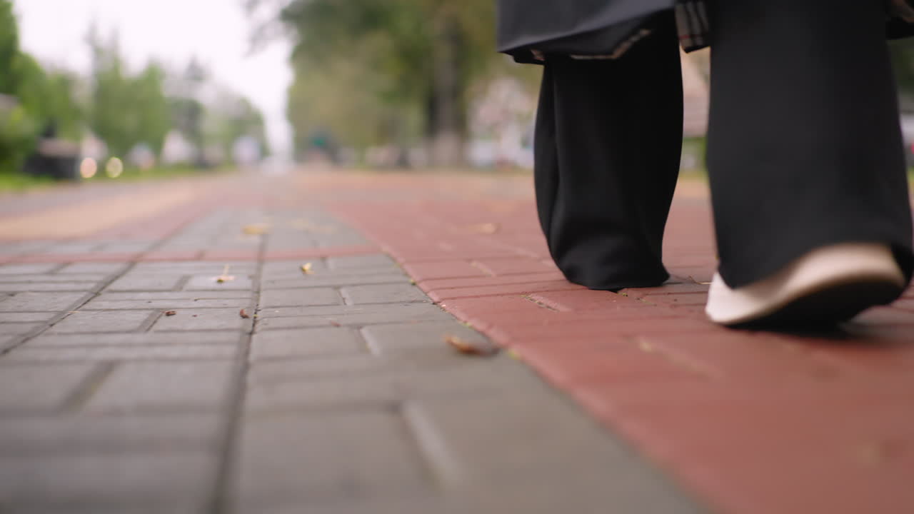 Close up of woman in black coat walking on brick pathway wearing sneakers, focus on step from behind with blurred cars and greenery in background, showing everyday lifestyle, autumn atmosphere and motion