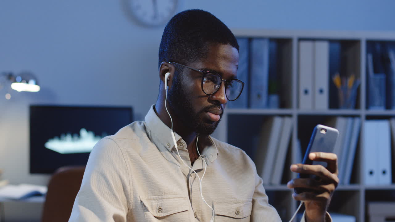 Close Up View Of Young Man In Glasses And Headphones Listening To The Music On Smartphone And Nodding Head In Rhythm In The Office At Night