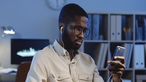 Vista-De-Cerca-De-Un-Joven-Con-Gafas-Y-Auriculares-Escuchando-Música-En-Un-Smartphone-Y-Asintiendo-Con-La-Cabeza-Al-Ritmo-En-La-Oficina-Por-La-Noche