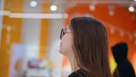 confident woman with long brown hair and glasses walks past vibrant store with an orange interior, she looks ahead with a relaxed expression, carrying a shopping bag in modern retail environment