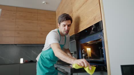 A-confident-brunette-cleaner-guy-with-stubble-in-a-white-T-shirt-and-a-blue-apron-washes-a-window-in-an-oven-using-a-yellow-rag-and-detergent-while-calling-a-cleaning-company-to-the-house