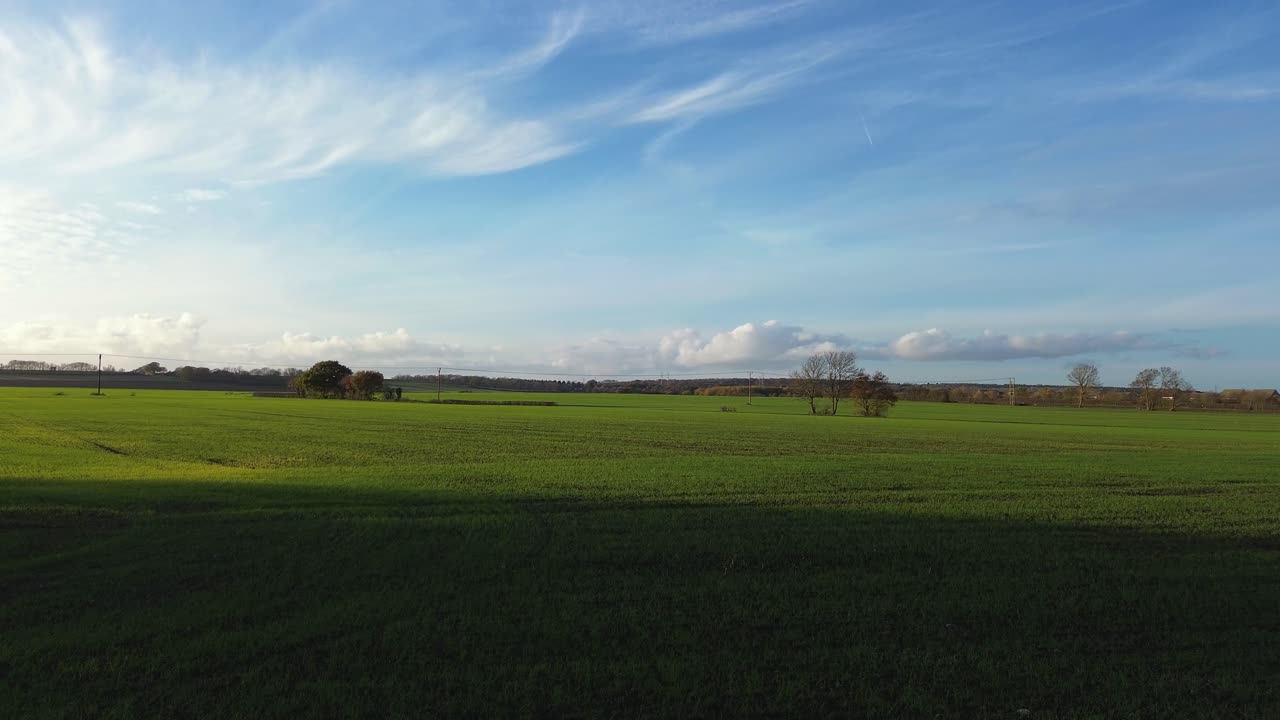 A serene aerial shot of the UK countryside, featuring lush green fields under a vibrant blue sky, with the sun setting in the distance and fluffy clouds scattered across the horizon
