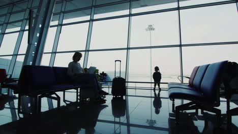 a woman with a child awaiting her flight sit on chairs in airport terminal silhouettes