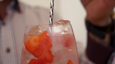 slowmo - male barman finishing gin and tonic drink coctail with strawberies and ice cubes in glass - close up