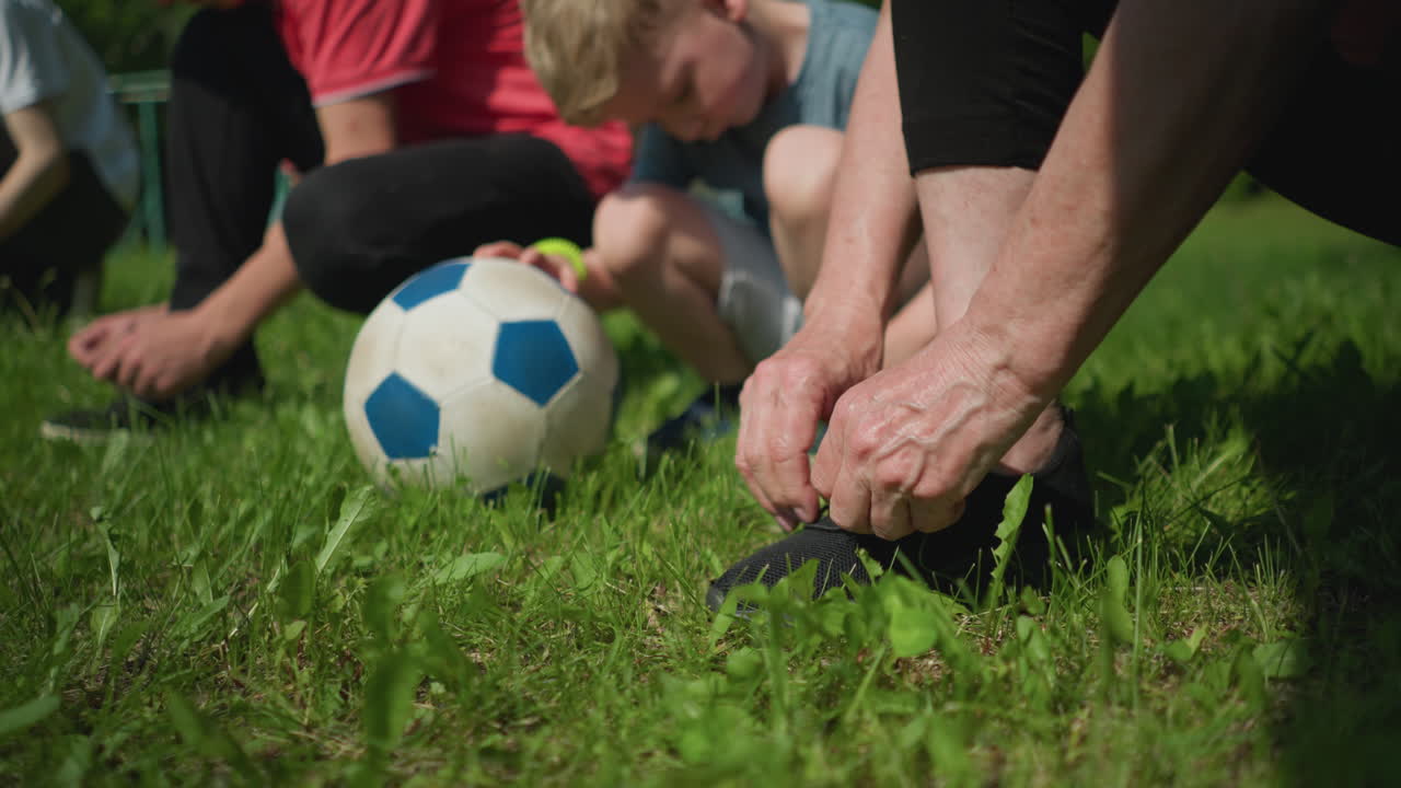 Close-up of four people in an outdoor setting, with three kneeling down to tie their shoelaces while a little boy places his hand on a soccer ball