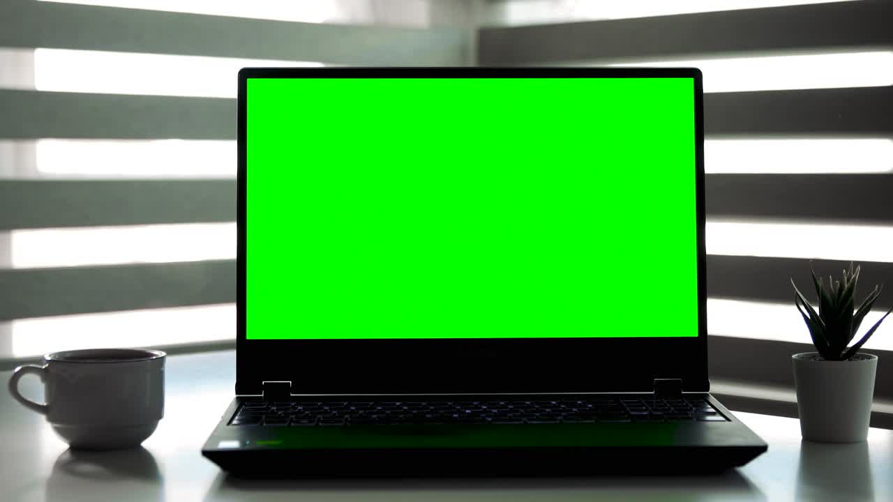 Laptop with green screen on white table in front of roller blind. With Coffee.