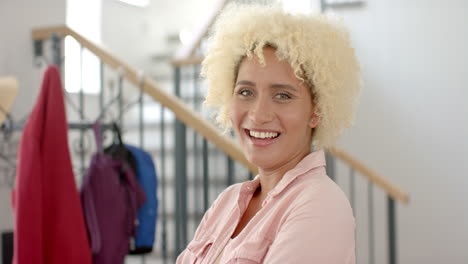 Young-biracial-woman-with-curly-blonde-hair-smiles-warmly-at-home,-wearing-a-pink-shirt