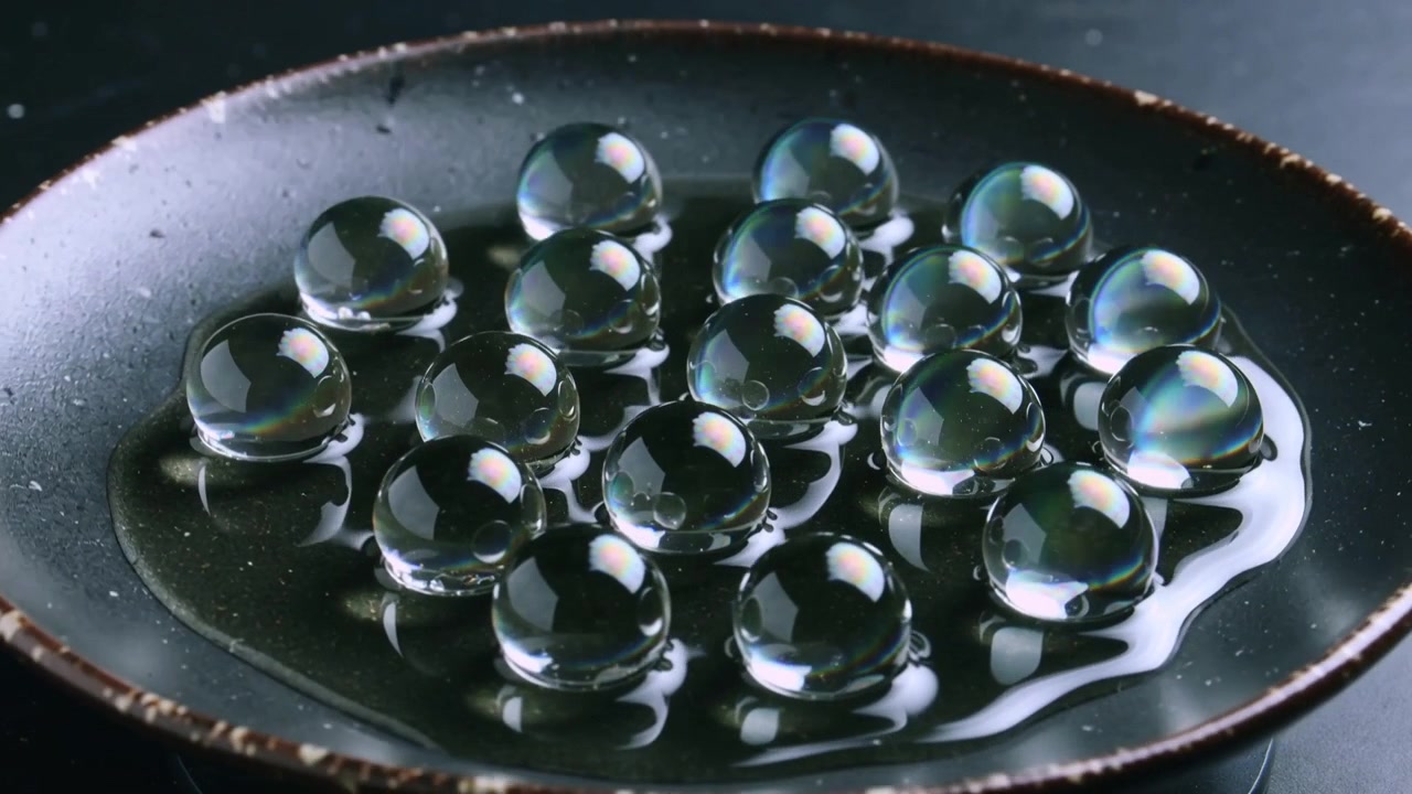 Illuminazione mutevole, gruppo di perline trasparenti che si posa su un piatto in ceramica scura poco profondo, liquido che increspa