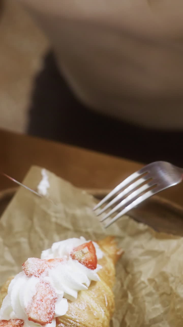 Nærbillede af en kvindehånd, der pakker en gaffel ud på en hyggelig café. Scenen viser en croissant med flødeskum og en kop kaffe på bordet. Afslappet atmosfære med blød baggrundsbelysning.