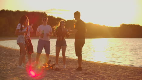 Die-Studenten-Tanzen-Bei-Sonnenuntergang-In-Shorts-Und-T-Shirts-Am-Sandstrand-Mit-Bier-Um-Das-Lagerfeuer.-Sie-Trinken-Bier-Und-Genießen-Den-Warmen-Sommerabend-Am-Fluss.