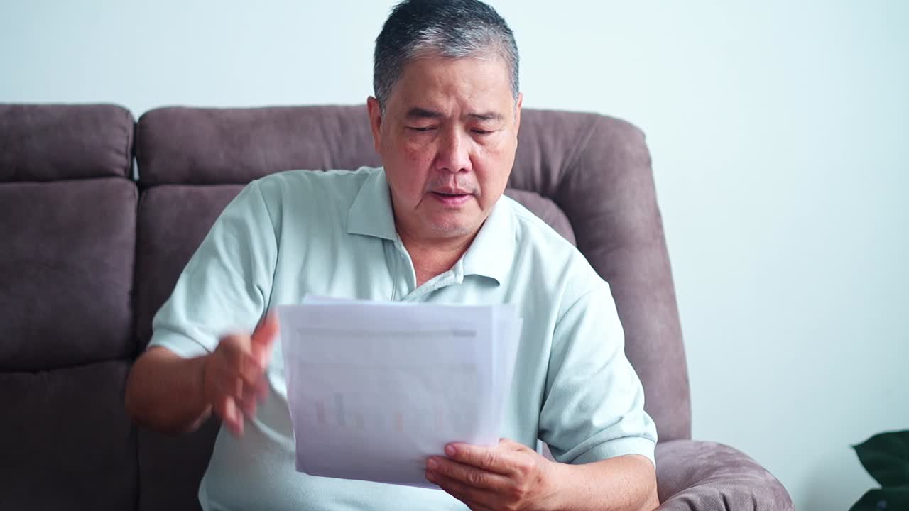 Elderly asian man stressed over bills while holding documents with worried expression at home
