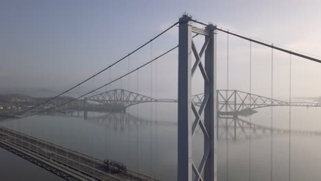 Aerial-footage-of-the-old-Forth-Road-Bridge-with-the-Forth-Railway-Bridge-in-the-background-on-a-sunny-day-at-South-Queensferry-in-West-Lothian,-Scotland