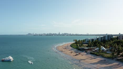 aerial drone wide shot of the tropical bessa beach in the capital city of joao pessoa, paraiba, brazil with people enjoying the ocean, a small tour boat, jet ski, and skyscrapers in the background