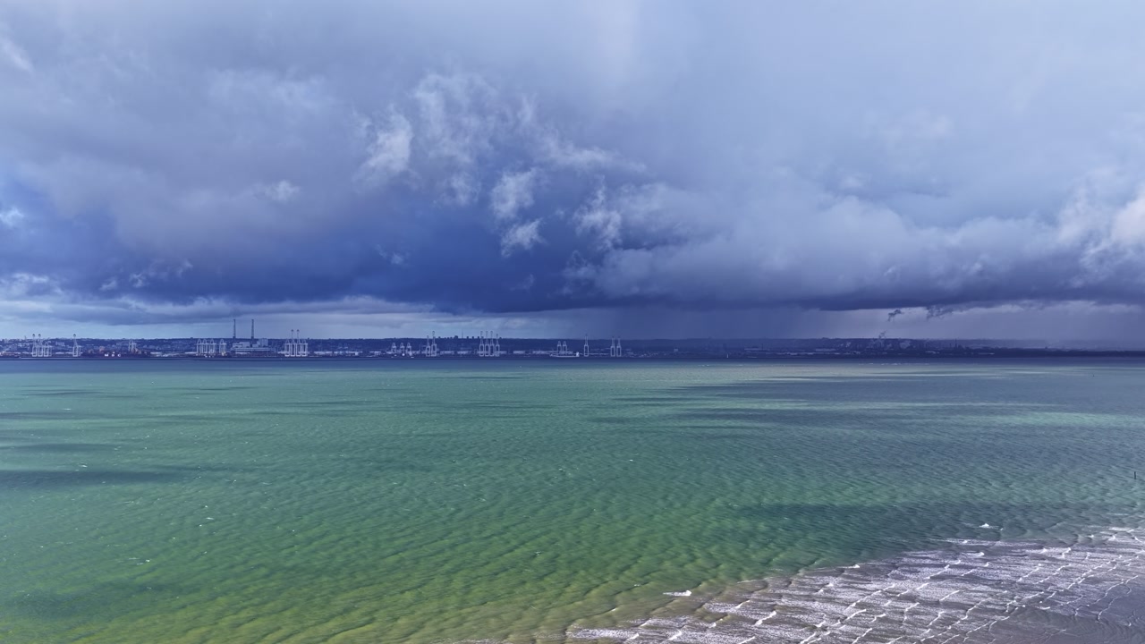 Dark storm clouds over open sea with distant industrial harbor skyline on horizon, aerial static timeapse of waves over buildings as clouds cast shadows on water