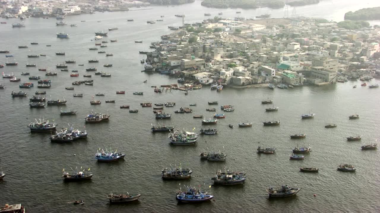 A dynamic aerial shot showcasing a bustling fishing harbor filled with tightly packed boats near a densely populated coastal town. The scene captures the rhythm of maritime life