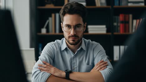 a focused young man with glasses and folded arms in an office setting, showcasing concentration and determination in a thoughtful moment