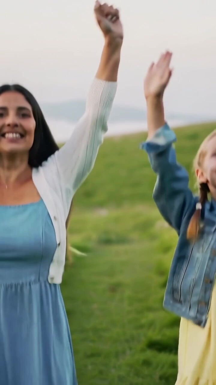 Video verticale: Ripresa dall'alto di mamma e figlia che alzano le braccia e saltano in riva al mare, indossando giacca di jeans.
