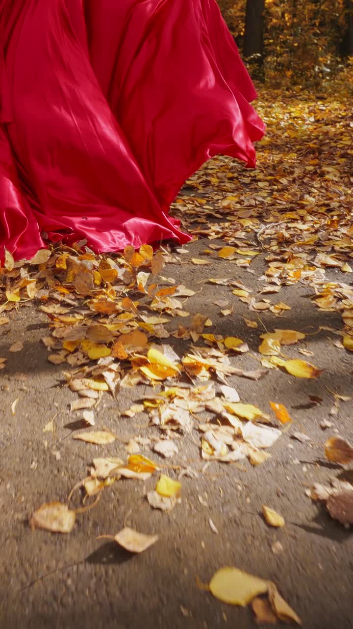 A Graceful Dance in Autumn: A Woman in a Flowing Red Dress Walks Through a Path Blanketed in Golden Leaves, Embracing the Beauty of Fall