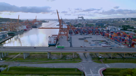 Puerto-De-Carga-De-Gdynia,-Grandes-Grúas-Cargando-Carga-En-Buques-Portacontenedores-Al-Atardecer,-Camión-De-Carga-Moviéndose-A-Lo-Largo-Del-Puente-Al-Atardecer