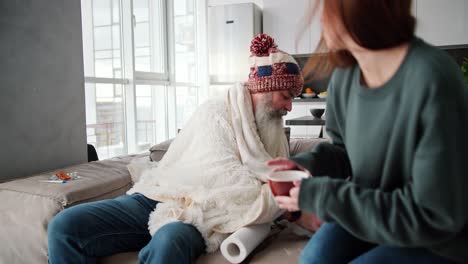 una chica morena con una chaqueta verde trae una taza de té a su padre enfermo de la vejez con una gruesa barba gris en un a cuadros blancos y lo cuida durante la enfermedad del hombre en un apartamento moderno