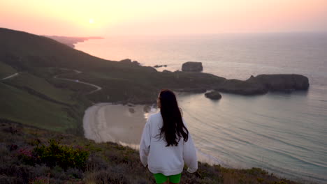 mujer joven con sudadera blanca con capucha parada en la cima de un acantilado ventoso con impresionantes vistas de las olas del océano al atardecer