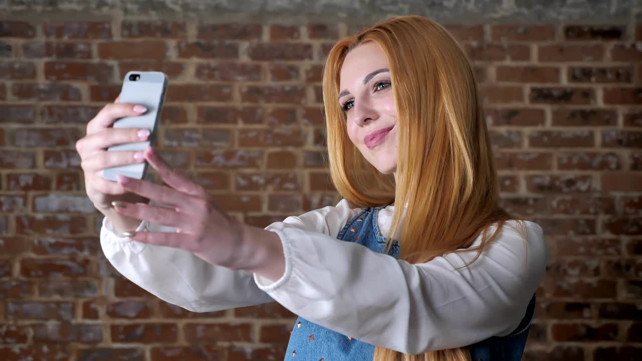 joven chica rubia dulce está haciendo selfie en el teléfono inteligente, sonriendo, fondo de ladrillo, concepto de comunicación