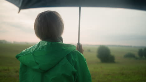 back view of middle-aged person in green raincoat standing in vast grassy field with an umbrella, looking into the distance under a cloudy sky, trees dot the far-off landscape