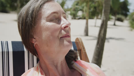una mujer caucásica mayor sonriente sentada en una silla de sol en una playa soleada con los ojos cerrados, en cámara lenta