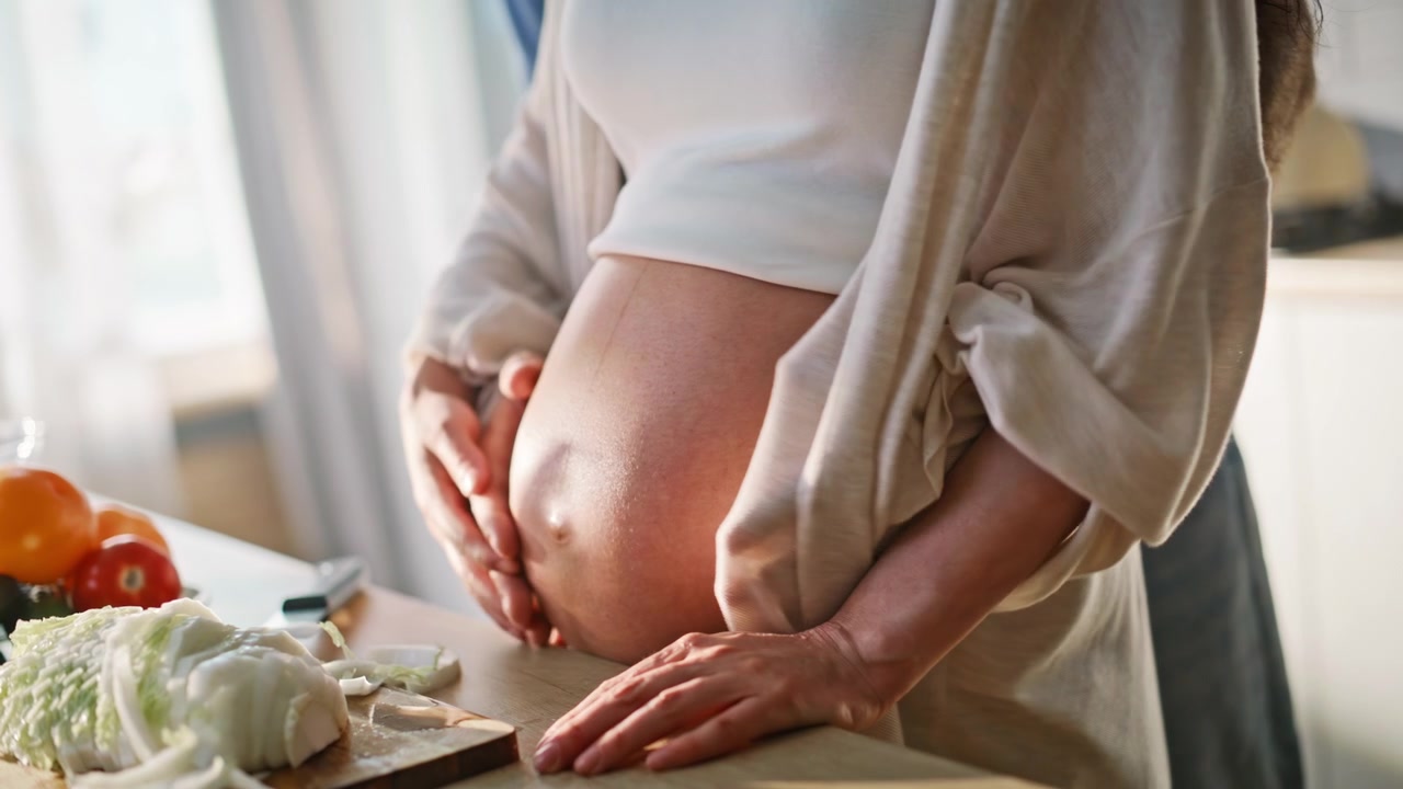 Ektemann omfavner gravid mage. Kvinne skjærer kål på skjærebrett på kjøkkenet.