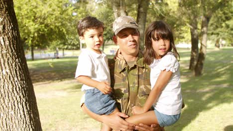 padre militar feliz sosteniendo a los niños en brazos al aire libre