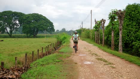 female riding bike on rocky dirt path in countryside away from camera, thailand