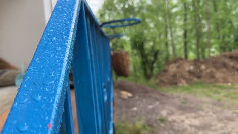 Ein-Ländliches-Dorf,-Malerische-Nahaufnahme-Von-Regentropfen-Auf-Einem-Blauen-Geländer,-Zeitlupe,-Grüner-Naturhintergrund-In-Einer-Ruhigen,-Entspannenden-Lodge-Auf-Dem-Land,-Reisekonzept-Im-Naturwandererholungstourismus