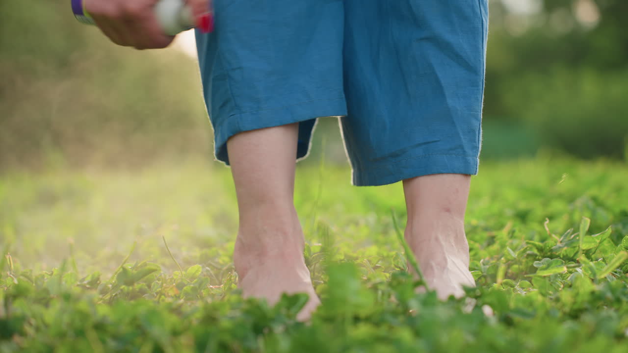 close up leg view of woman standing barefoot on green grass spraying insect mist on leg in warm evening light outdoor protection from bugs and wellness routine in nature park setting