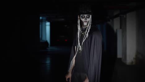 Creepy-man-with-skeleton-makeup-walking-on-dark-parking-hallway,-looking-at-camera,-trying-to-scare