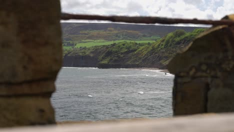 lookout point through stone wall fence showing waves crashing towards the shore with large rocky cliffs in the background on sunny day at north yorkshire coast